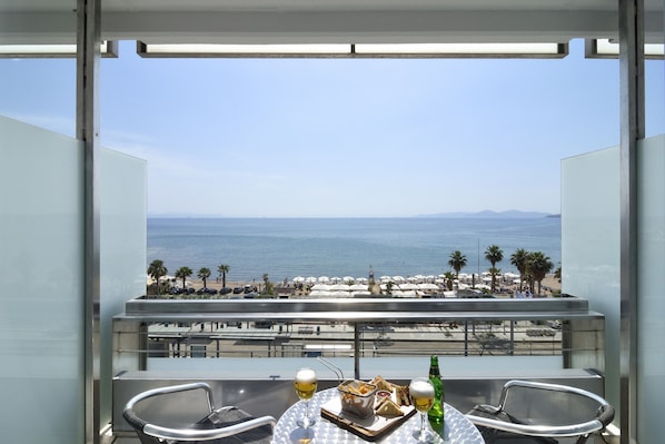 Un balcón con mesa lista para dos personas, con vista a una playa con sombrillas y palmeras.
