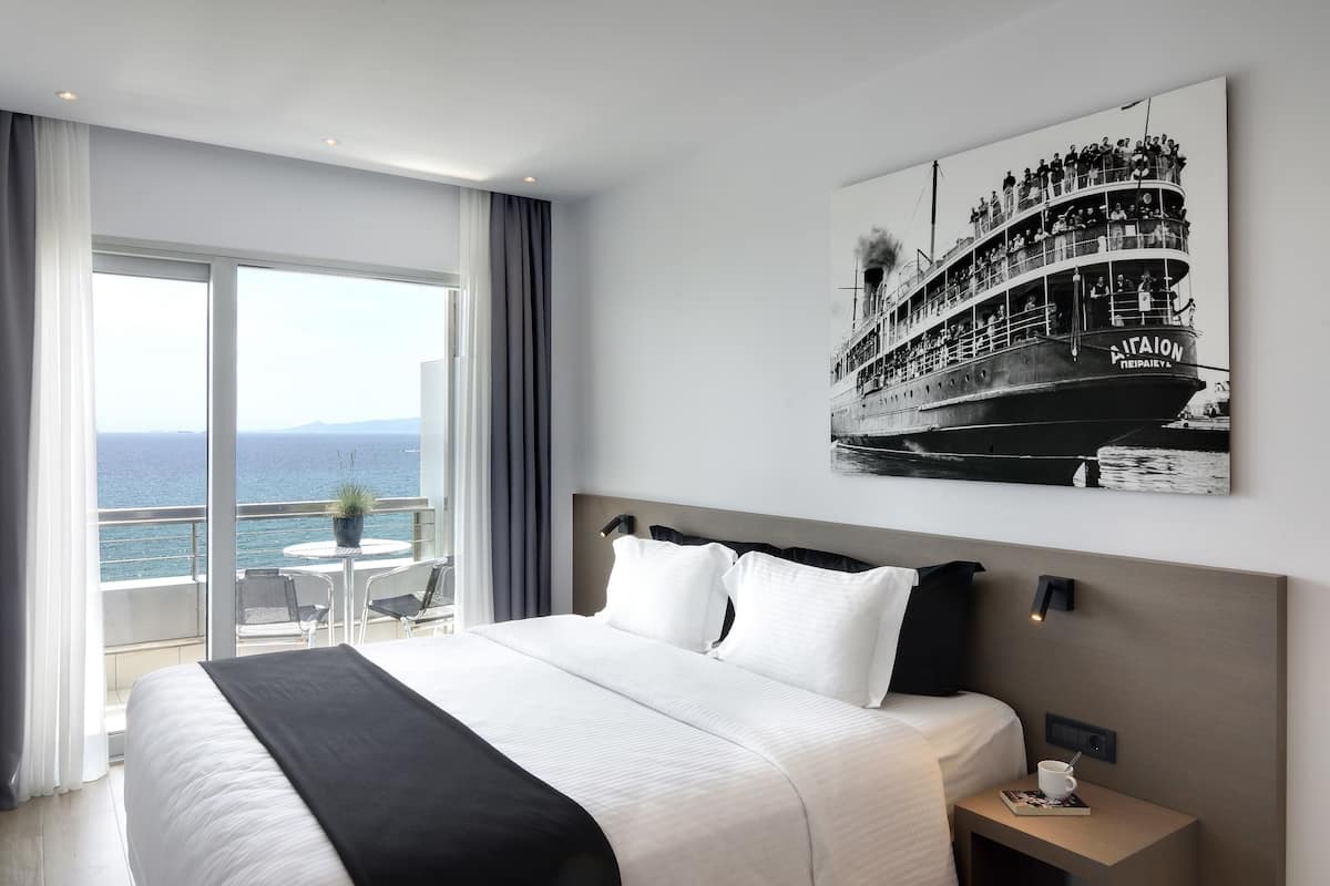 A hotel room with a large bed, a view of the sea, a balcony with a table and chairs, and a painting of a boat on the wall.