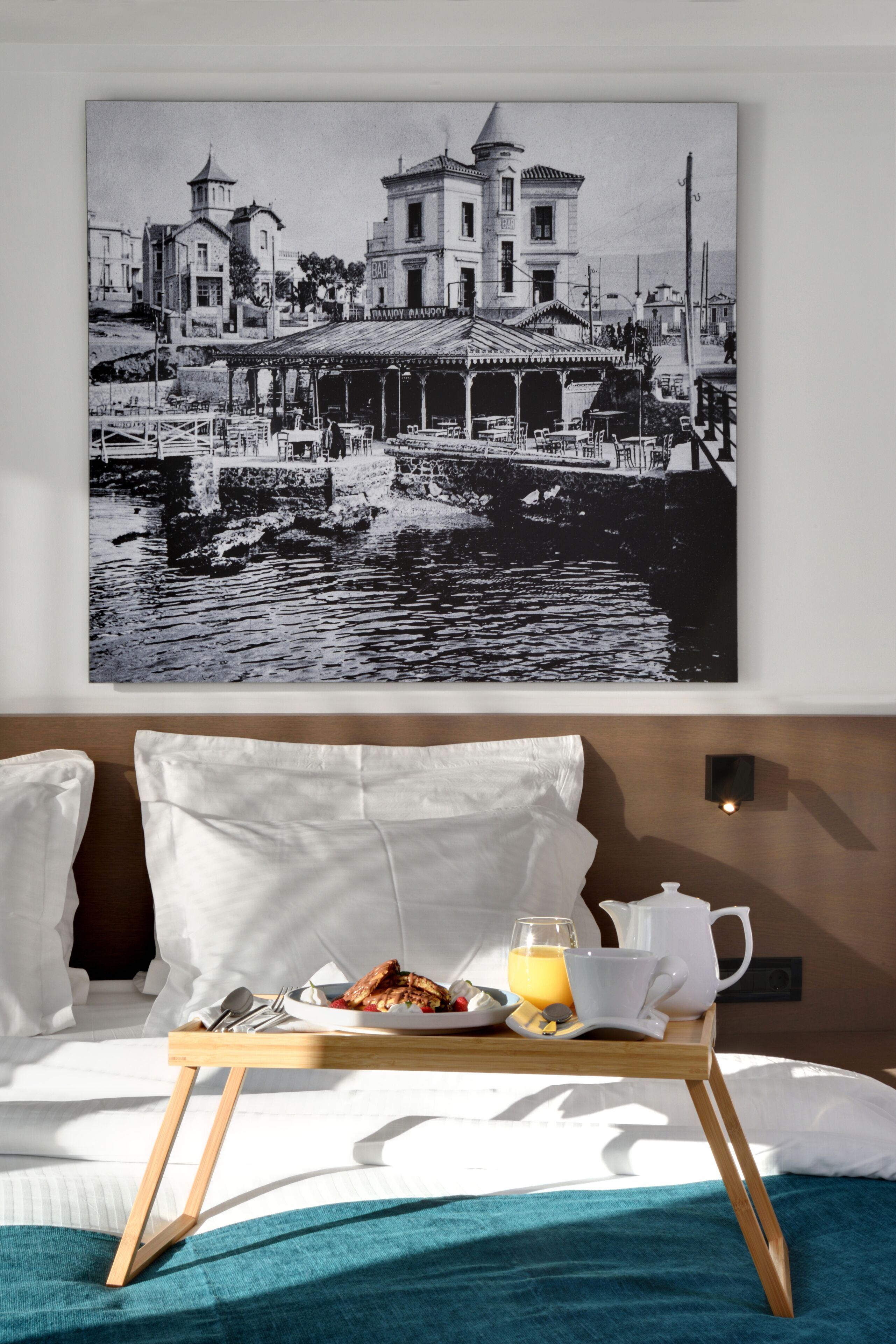 A hotel room with a bed, a breakfast tray with food and drinks, and a large black and white photo of a waterfront scene on the wall.