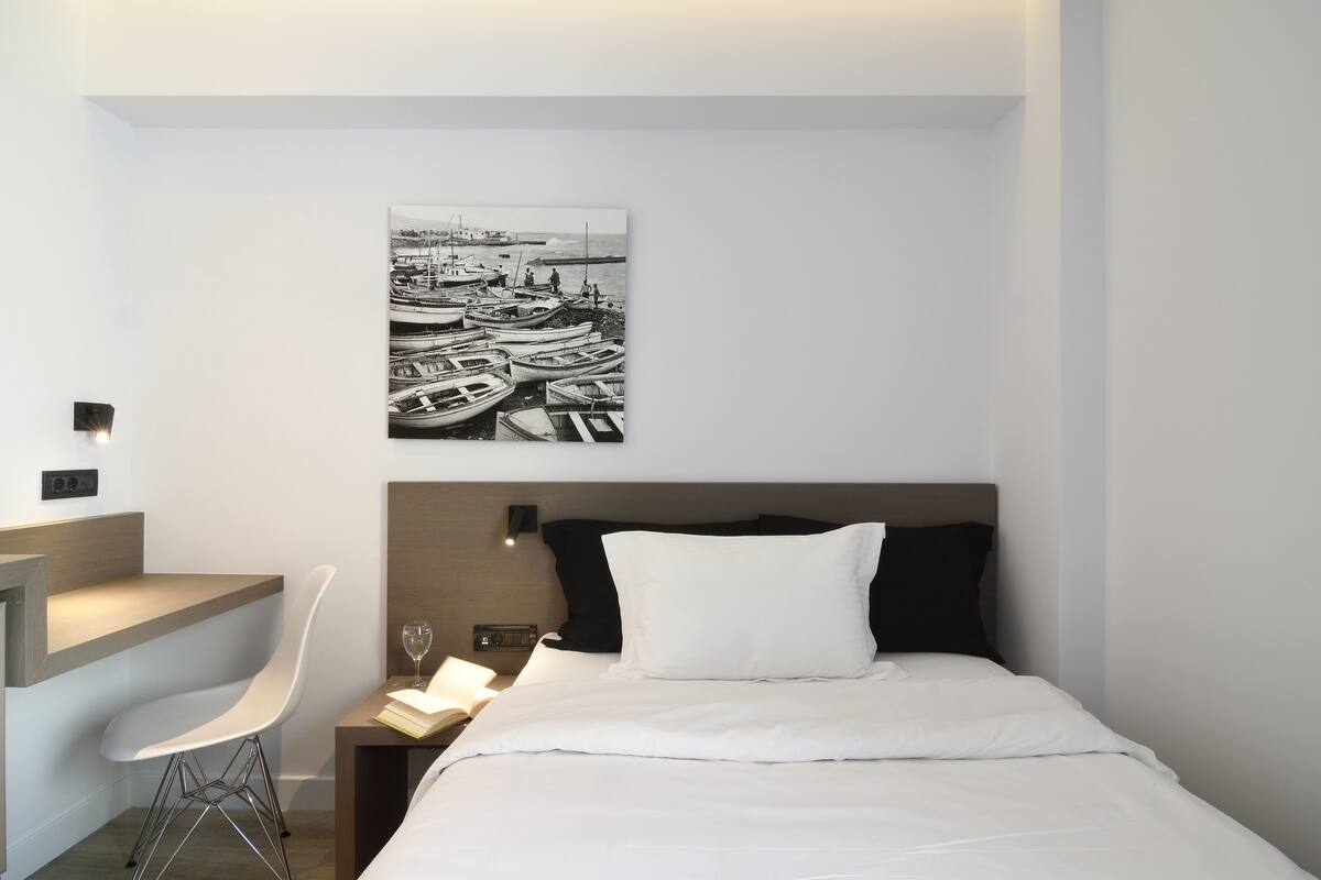 A modern hotel room with a bed, a desk, a chair, and a wall-mounted picture of boats.