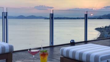 Varanda com vista para o mar e para a cidade, com uma mesa, duas bebidas e almofadas listradas.