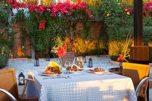 An outdoor dining area with a table set for a meal, surrounded by wicker chairs and a backdrop of flowering plants and a rustic wall. - Poseidon Athens (Palaio Faliro)