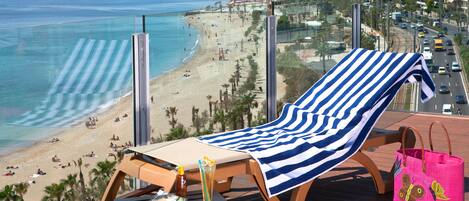 Un balcon avec vue sur la plage, une chaise longue, une table avec une boisson et un sac rose.