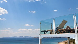 A glass-enclosed lounge area with two lounge chairs and a table, overlooking a beach and the sea.