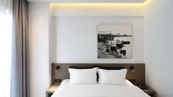 Una habitación de hotel moderna con una cama bien hecha, un cabecero con iluminación integrada, una mesita de noche con teléfono y una obra de arte en la pared que representa una escena costera.