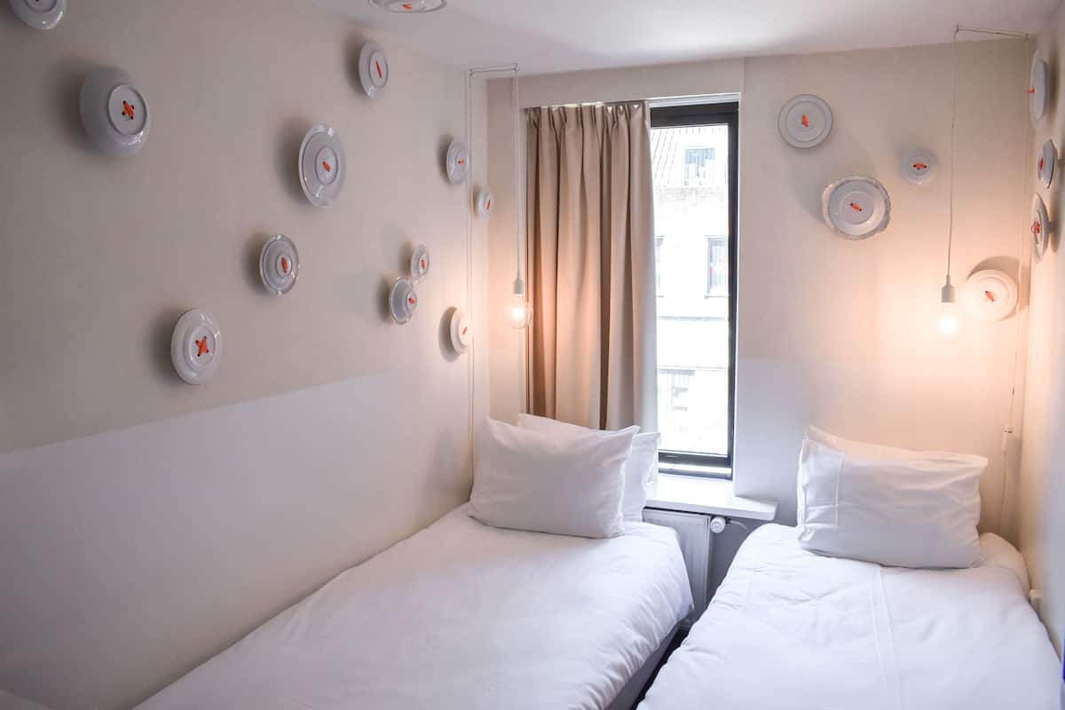 A hotel room with two beds, a window with curtains, and wall-mounted light fixtures.