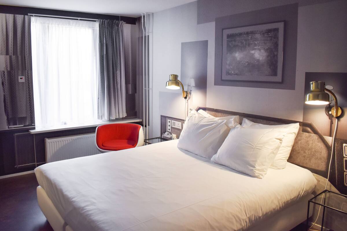 A hotel room with a large bed, two bedside lamps, a red chair, a window with curtains, and a framed picture on the wall.