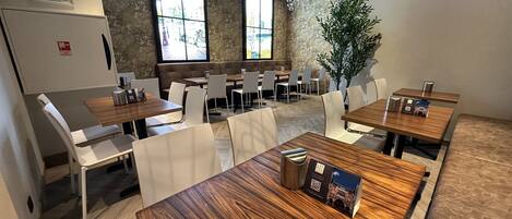 A modern dining area with wooden tables and white chairs, large windows, and a stone wall.