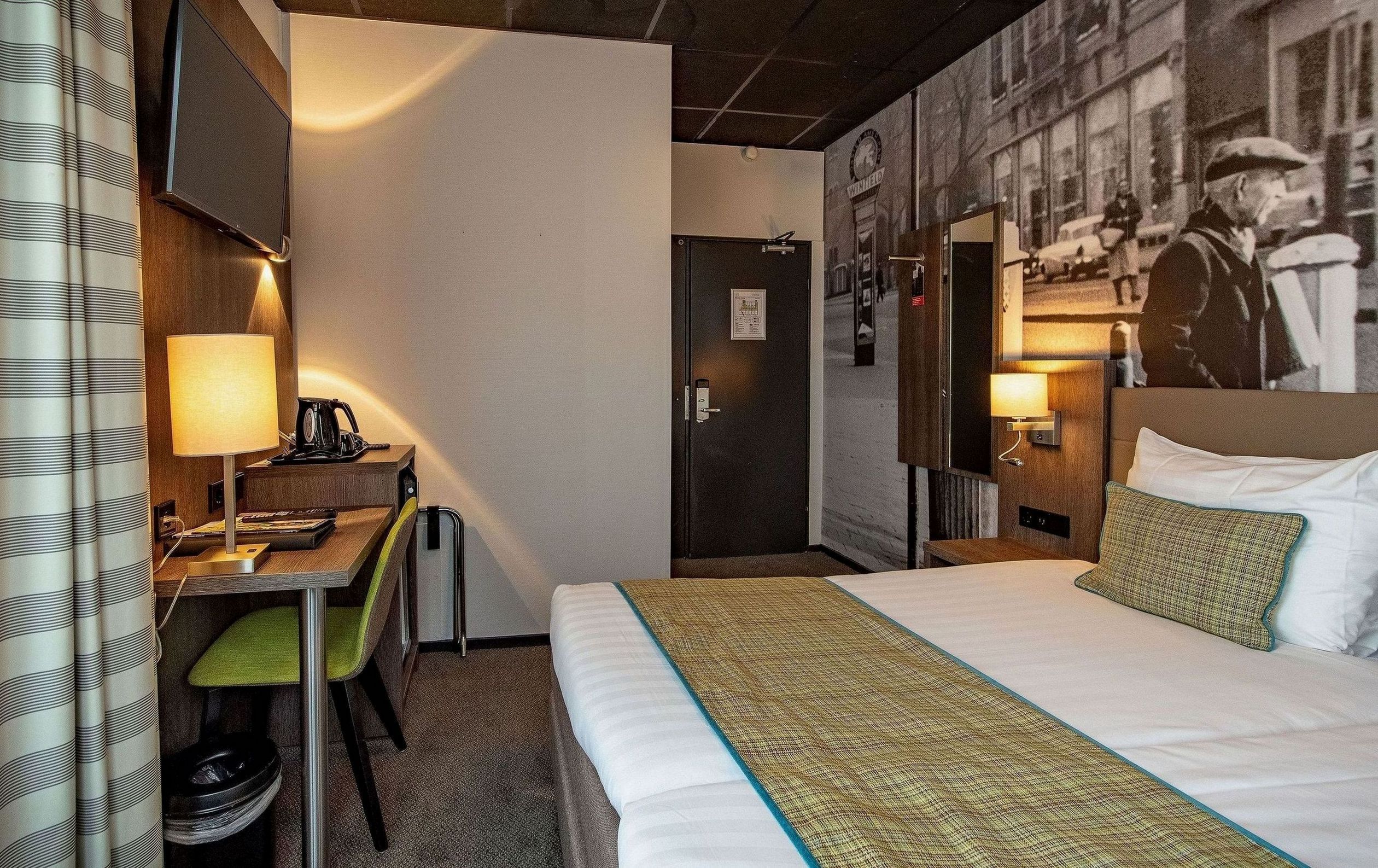 Habitación de hotel con cama, escritorio con lámpara, una silla y una pared con una foto antigua.