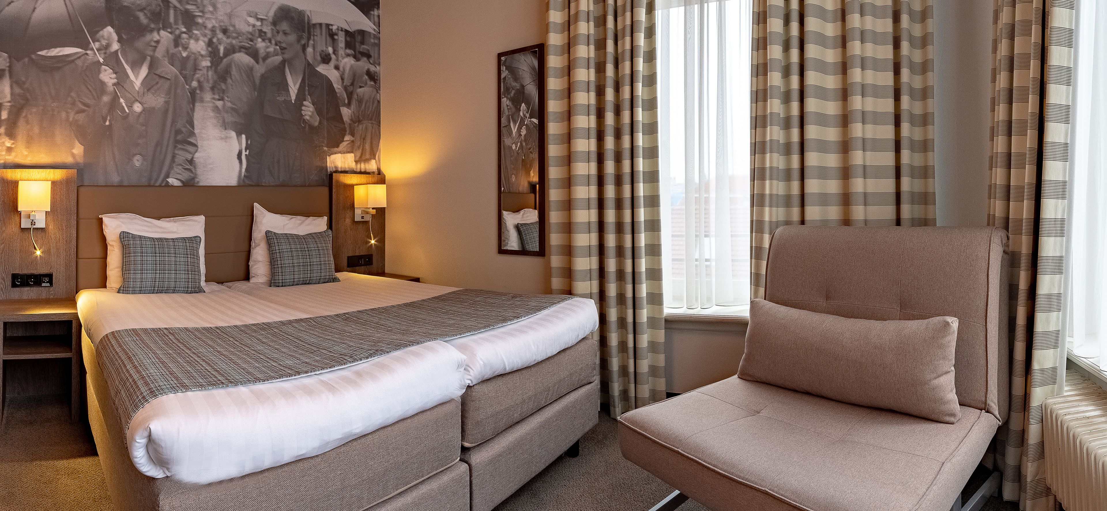 Una habitación de hotel con una cama grande, una silla, una mesita de noche y una lámpara fijada a la pared.