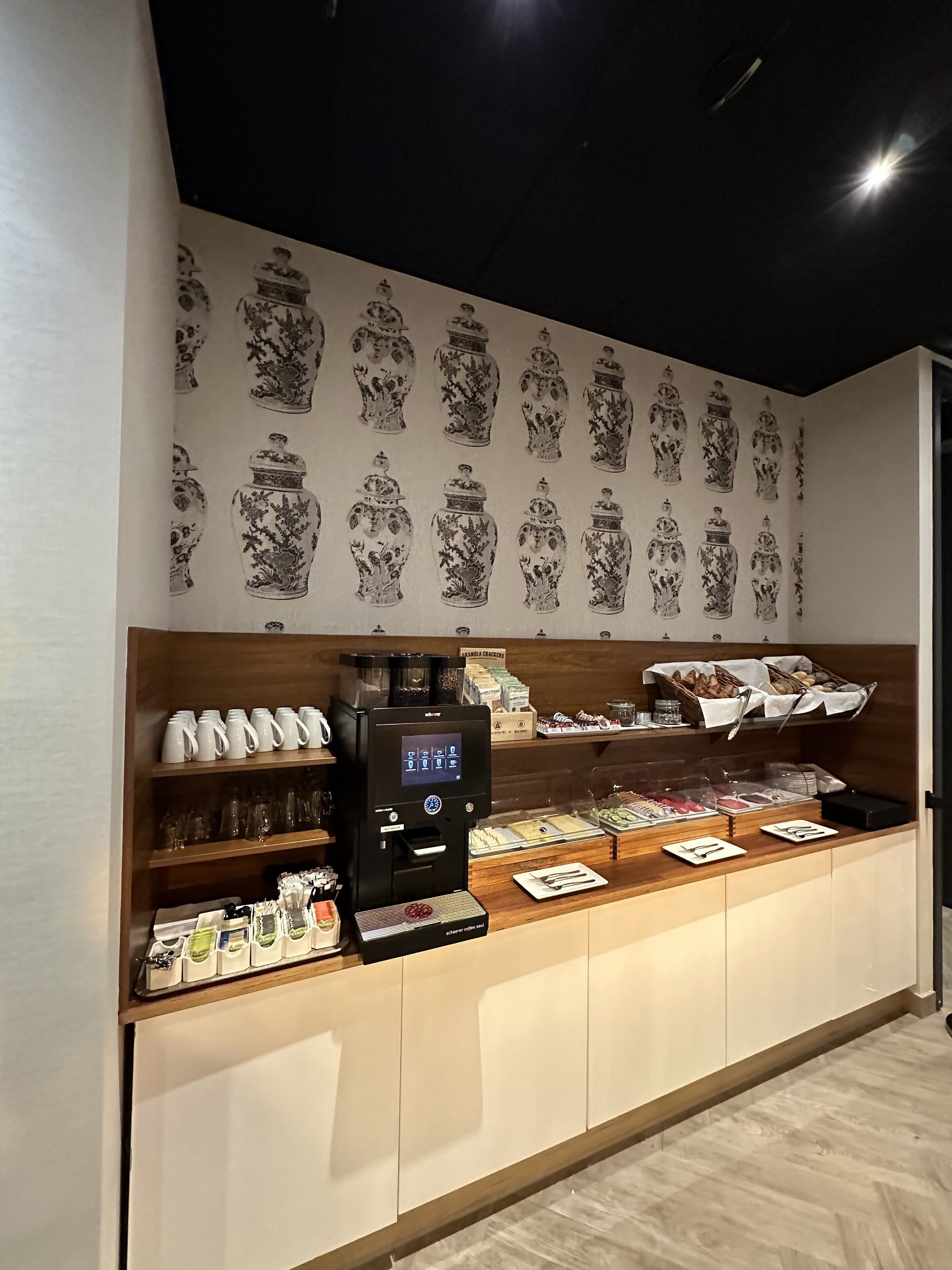 Una barra de desayuno con una cafetera, diversos alimentos y jarrones decorativos en la pared.