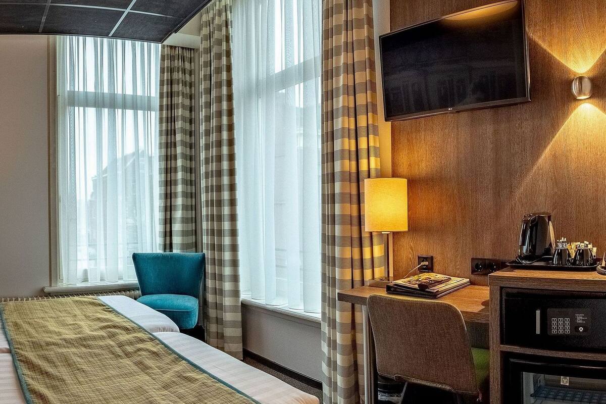 A hotel room with a bed, a desk with a computer, a TV, and a window with curtains.