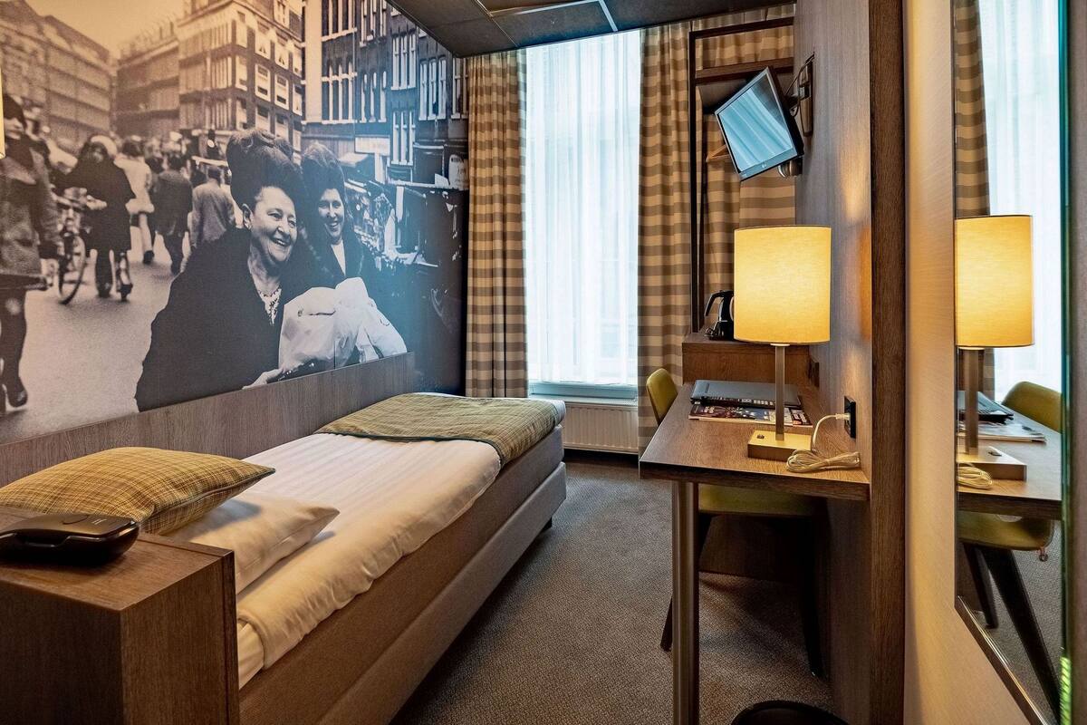 A hotel room with a bed, a desk, a chair, a TV, and a wall with a mural.