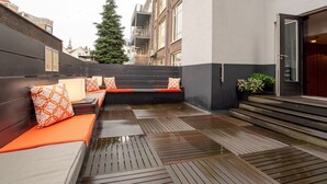 A covered outdoor seating area with wooden benches and orange cushions, a potted plant, and a view of the cityscape.