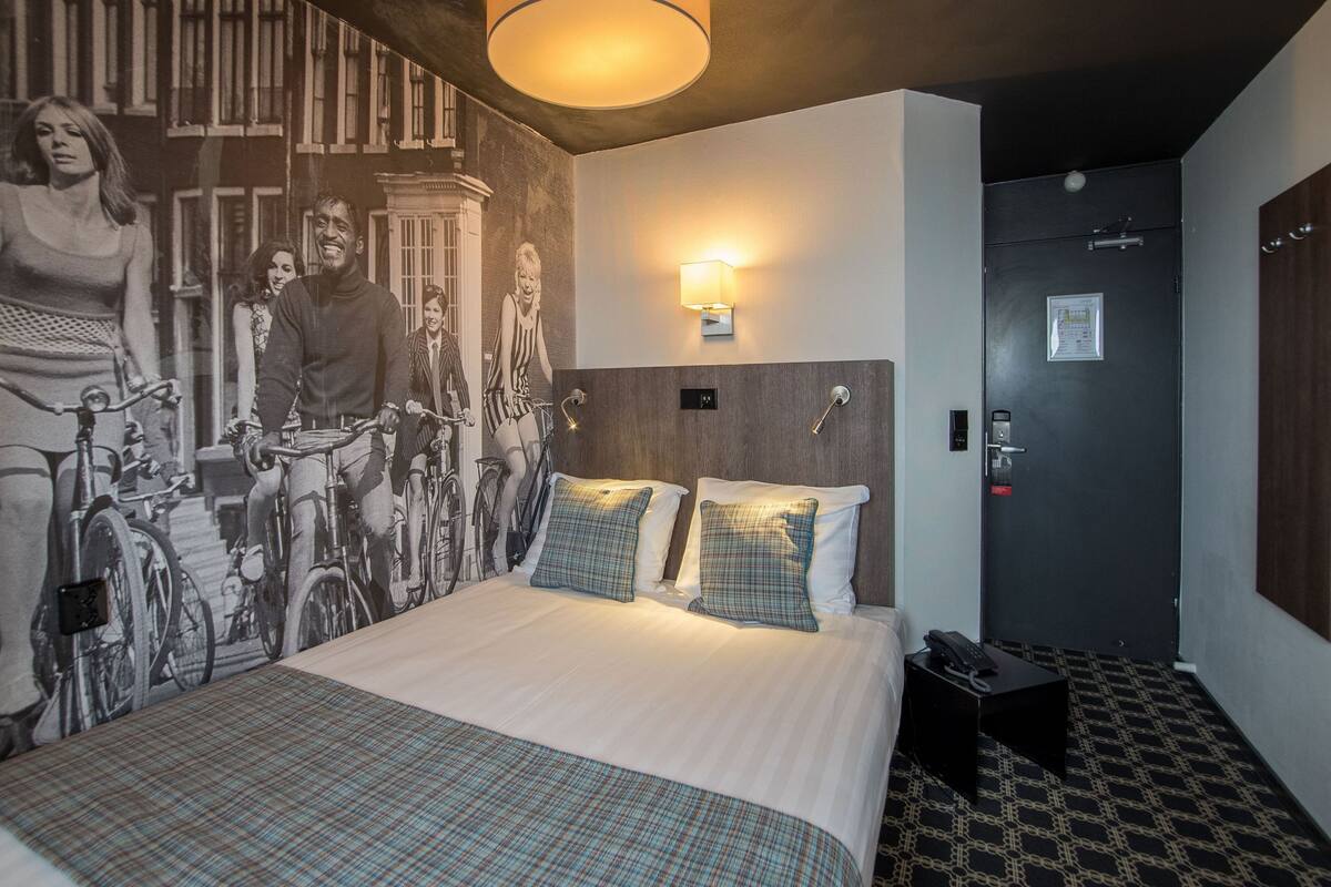 A hotel room with a large bed, a wall mural, a wall-mounted light, and a door with a lock.