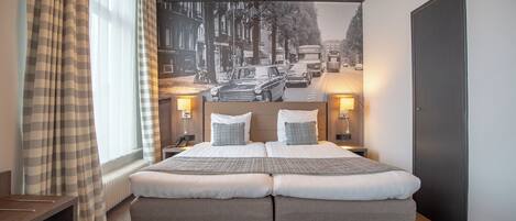 A double bed with two pillows, a nightstand with a lamp, a window with curtains, and a wall mural of a street scene.