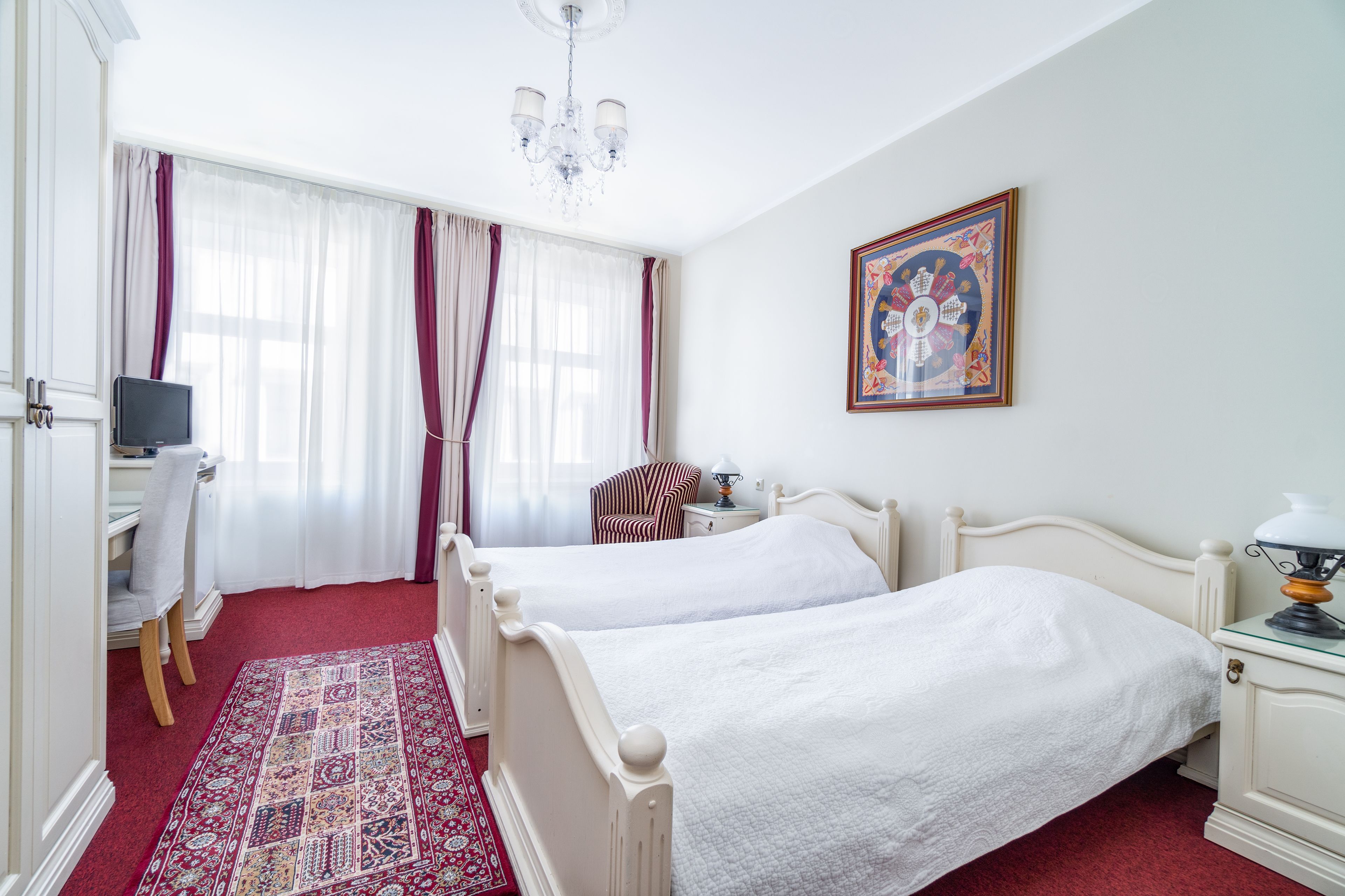 Hotellihuone, jossa on kaksi sänkyä, työpöytä tuolilla, televisio, kuviollinen matto ja kattokruunu.