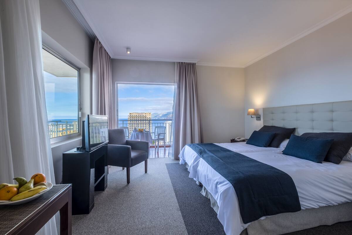 Hotelkamer met een groot bed, een televisie, een bureau met fruit en uitzicht op de stad.