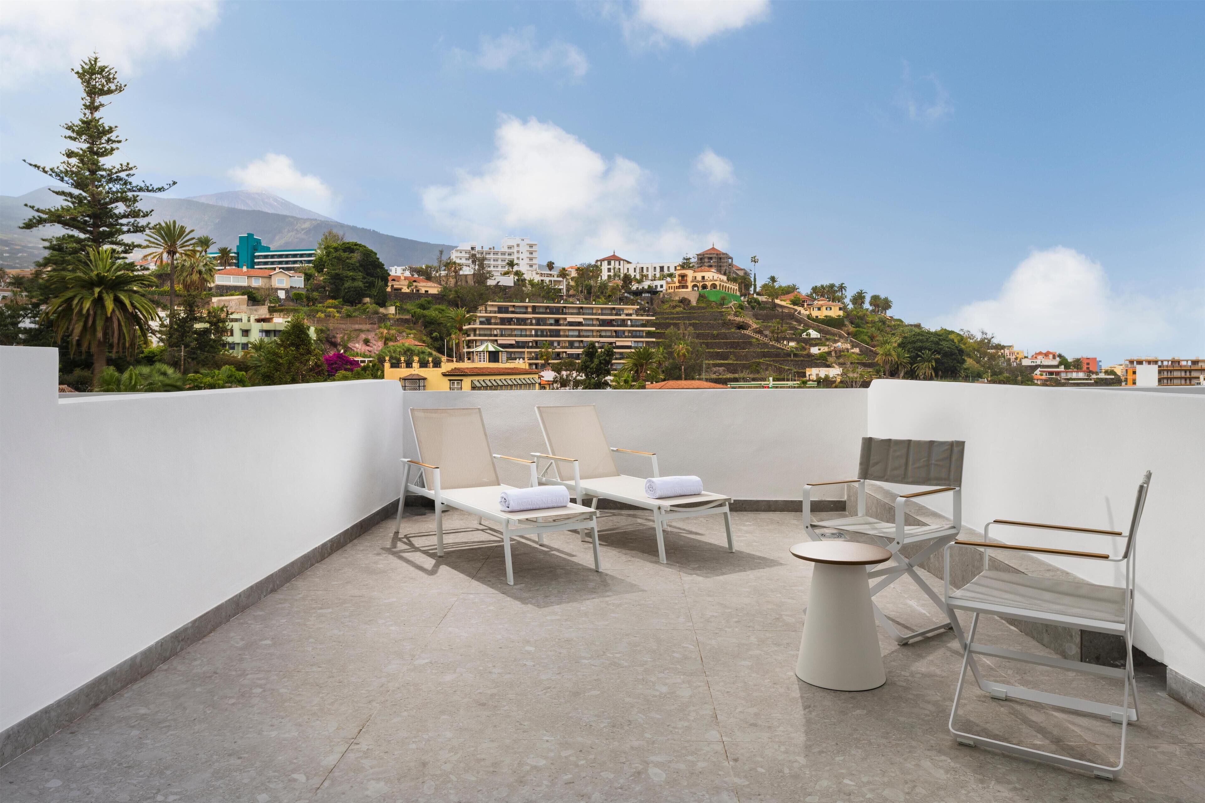 En tagterrasse med hvide metalstole og et rundt bord, med udsigt over en bakke med bygninger og et bjerg i baggrunden.