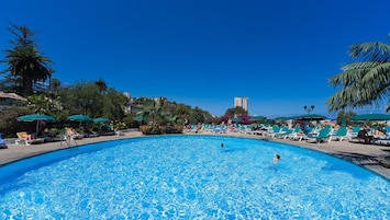 Una gran piscina al aire libre rodeada de sillas de descanso y sombrillas, con palmeras y edificios al fondo.