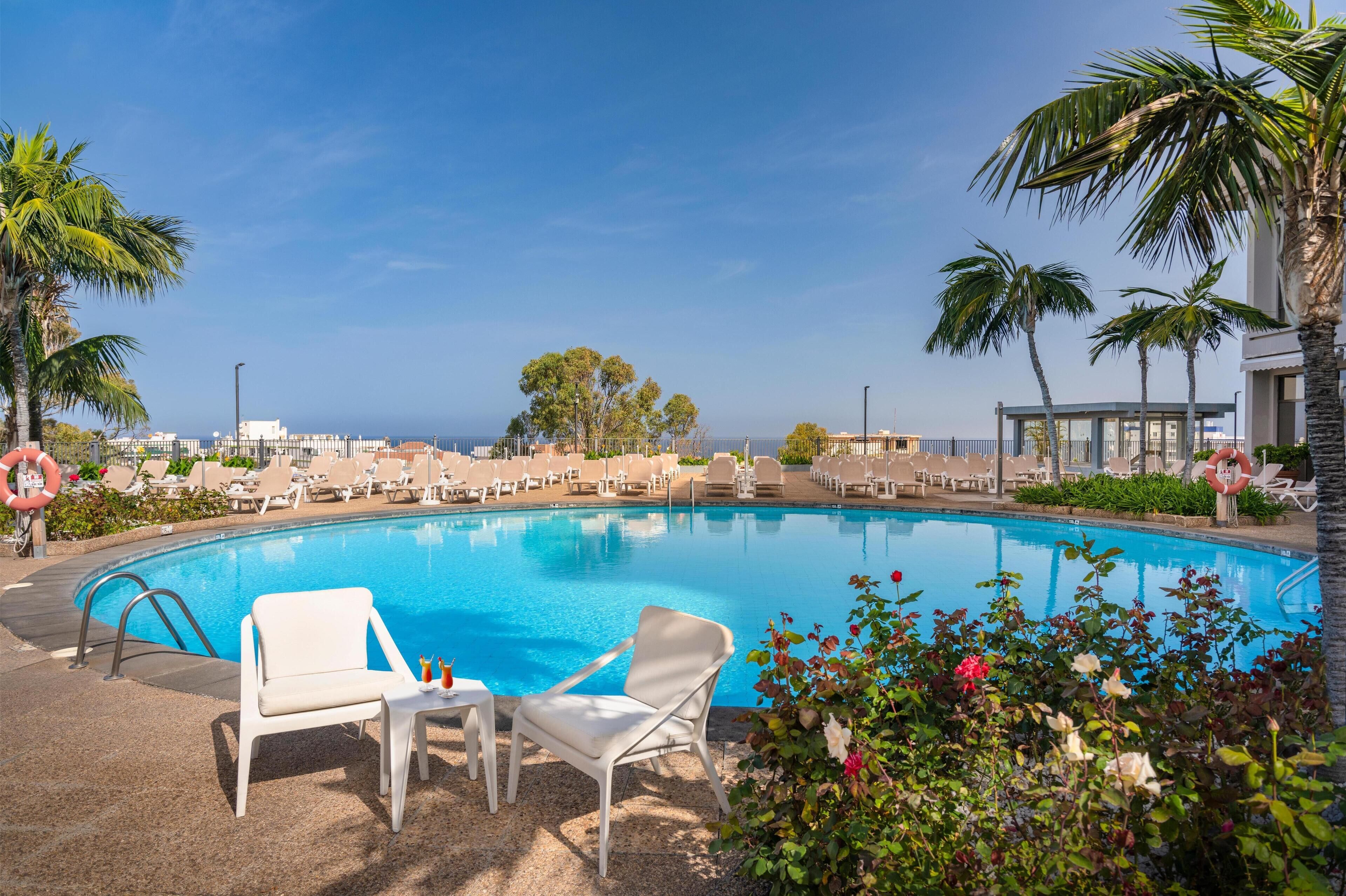 A pool area with lounge chairs and a lifebuoy, surrounded by palm trees and a clear sky.