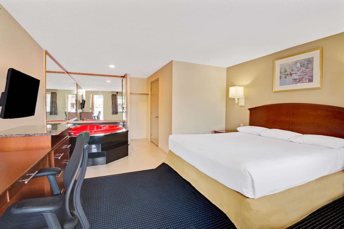 A hotel room with a bed, a desk with a chair, a mirror, a red sink, and a painting on the wall.