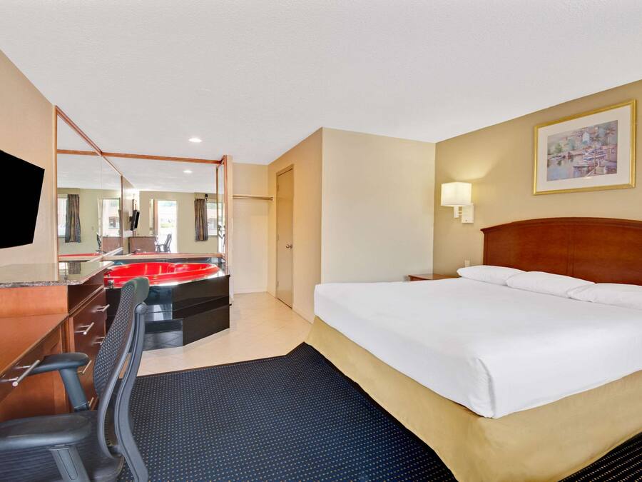Habitación de hotel con cama, escritorio con silla, espejo, lavamanos rojo y un cuadro en la pared.