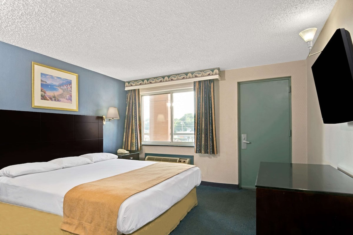 A hotel room with a bed, a desk, a lamp, a TV, a window with curtains, and a framed picture on the wall.