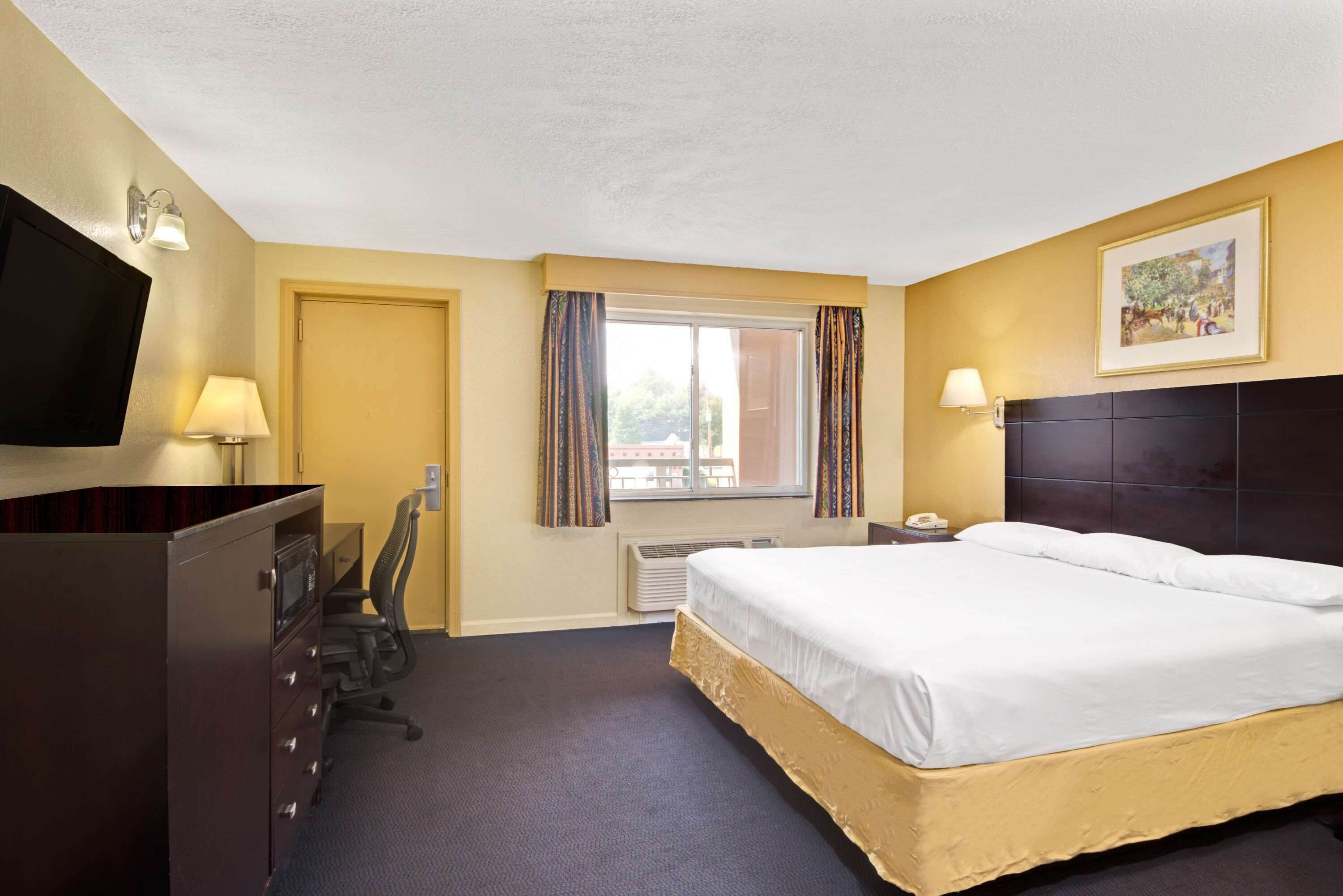 Een hotelkamer met een groot bed, een bureau met een stoel, een televisie, een raam met gordijnen en een schilderij aan de muur.