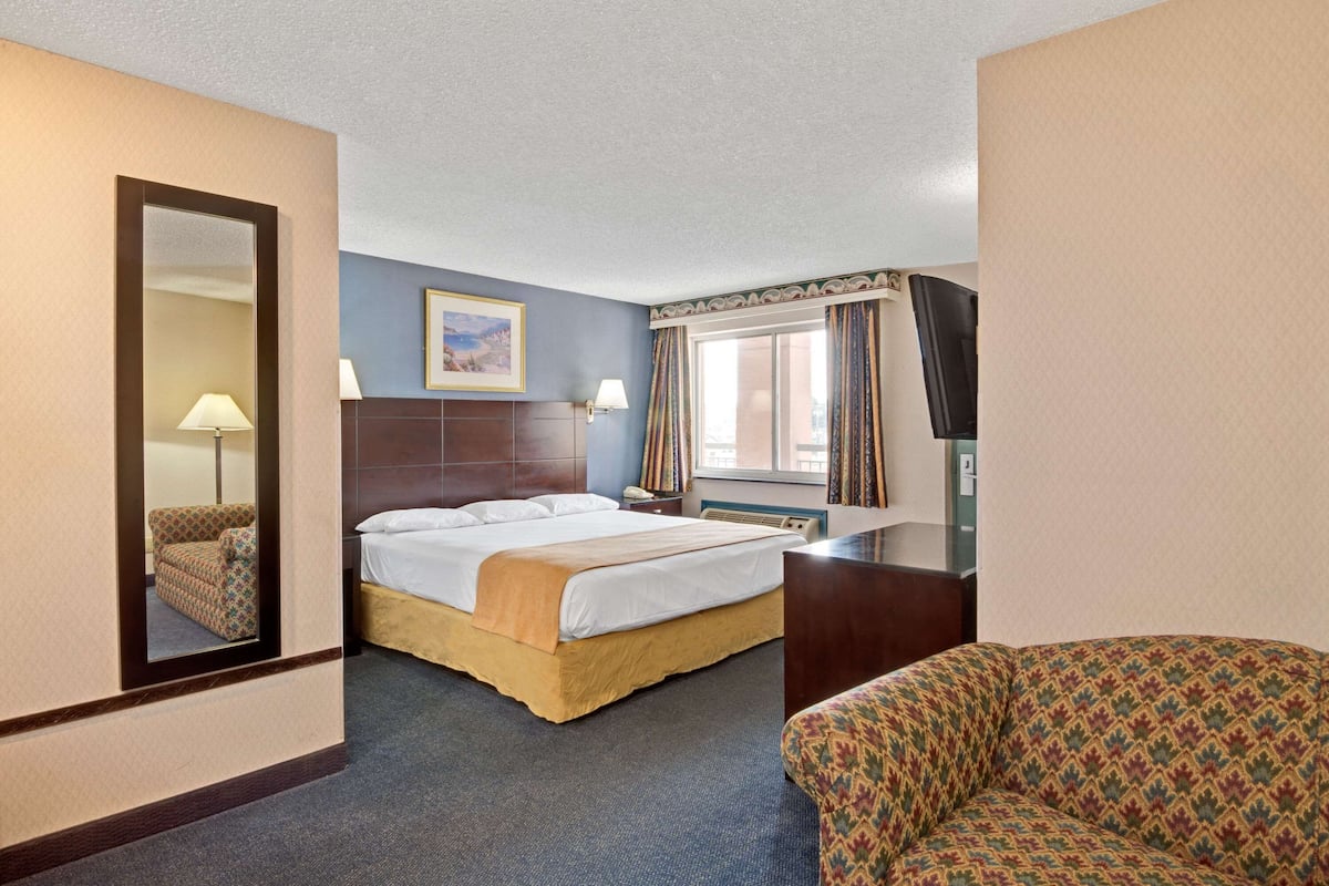 A hotel room with a bed, a chair, a desk, a mirror, a window with curtains, and a painting on the wall.