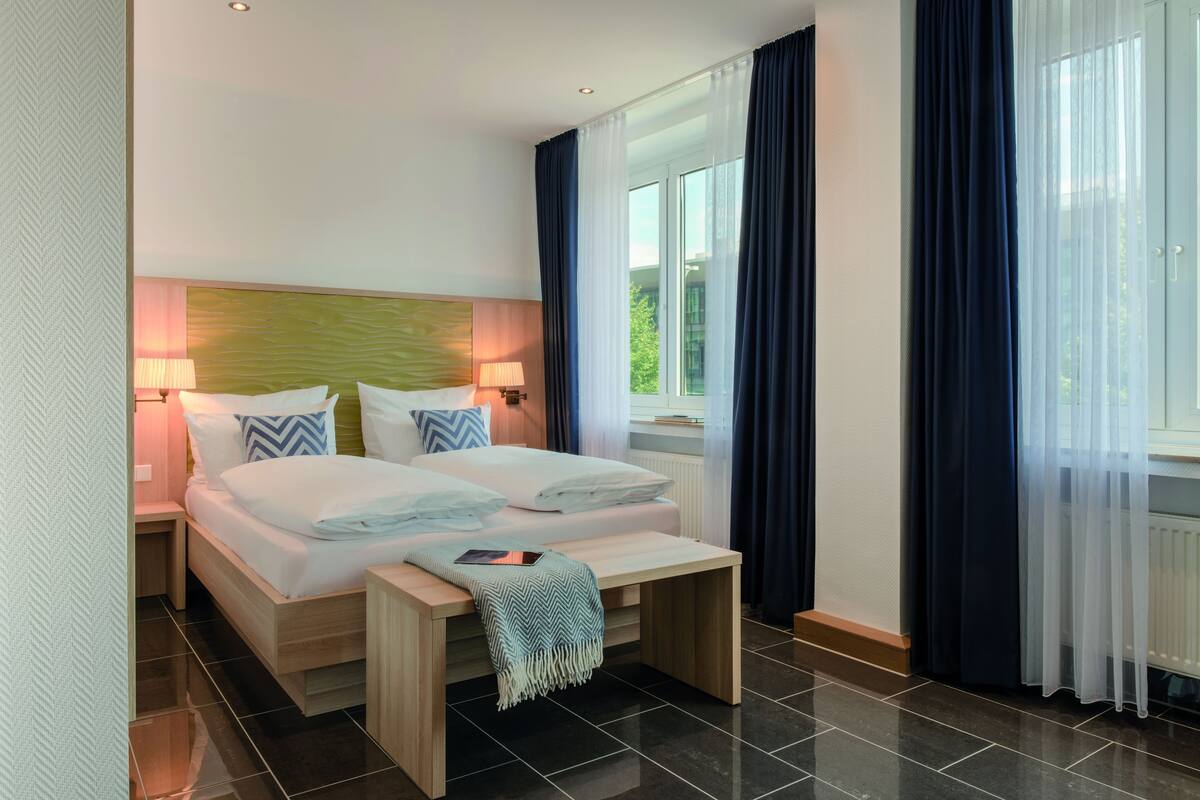 A hotel room with a large bed, bedside tables, a bench, and a view of greenery through the window.