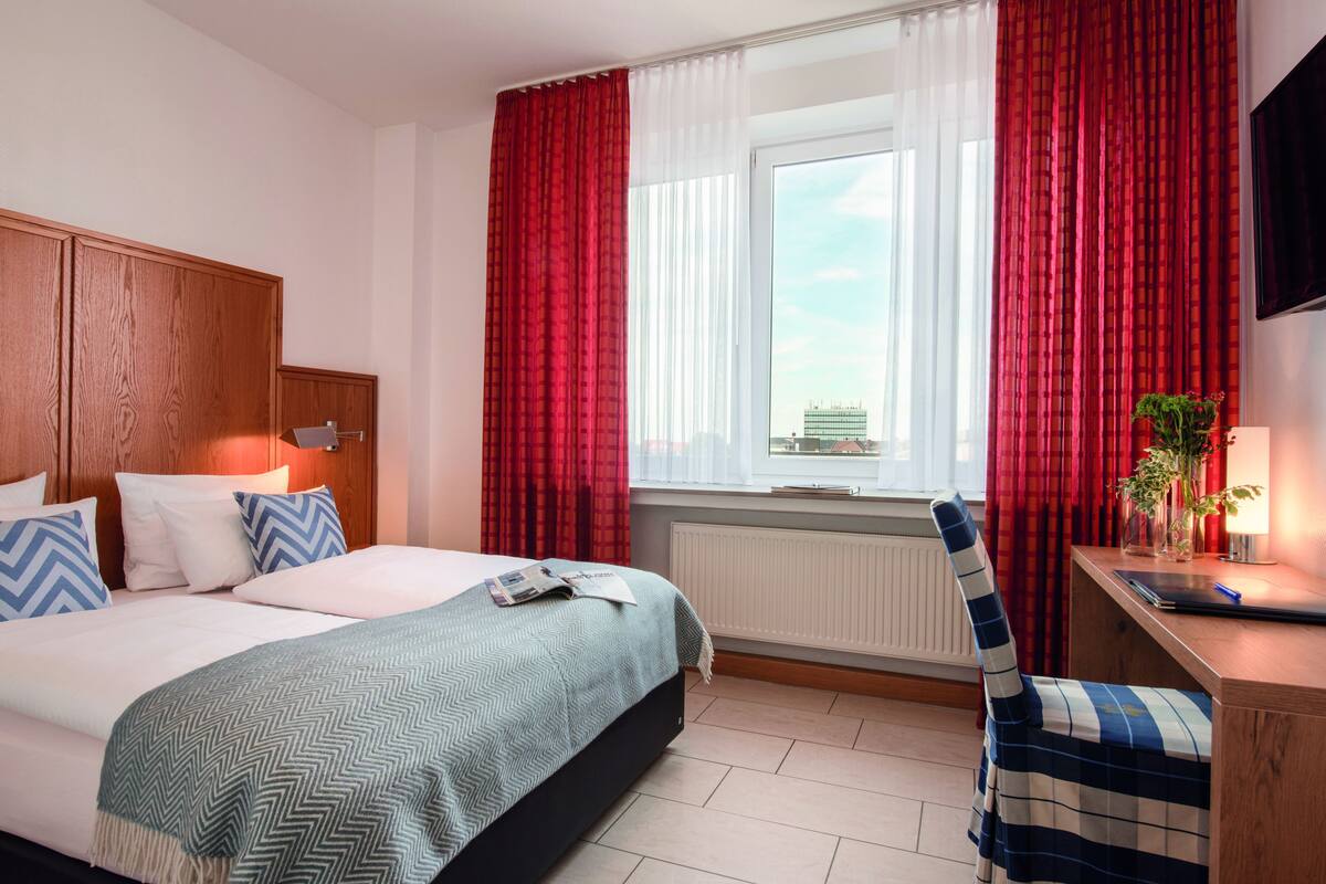A hotel room with a bed, a desk, a chair, and a window with red curtains.