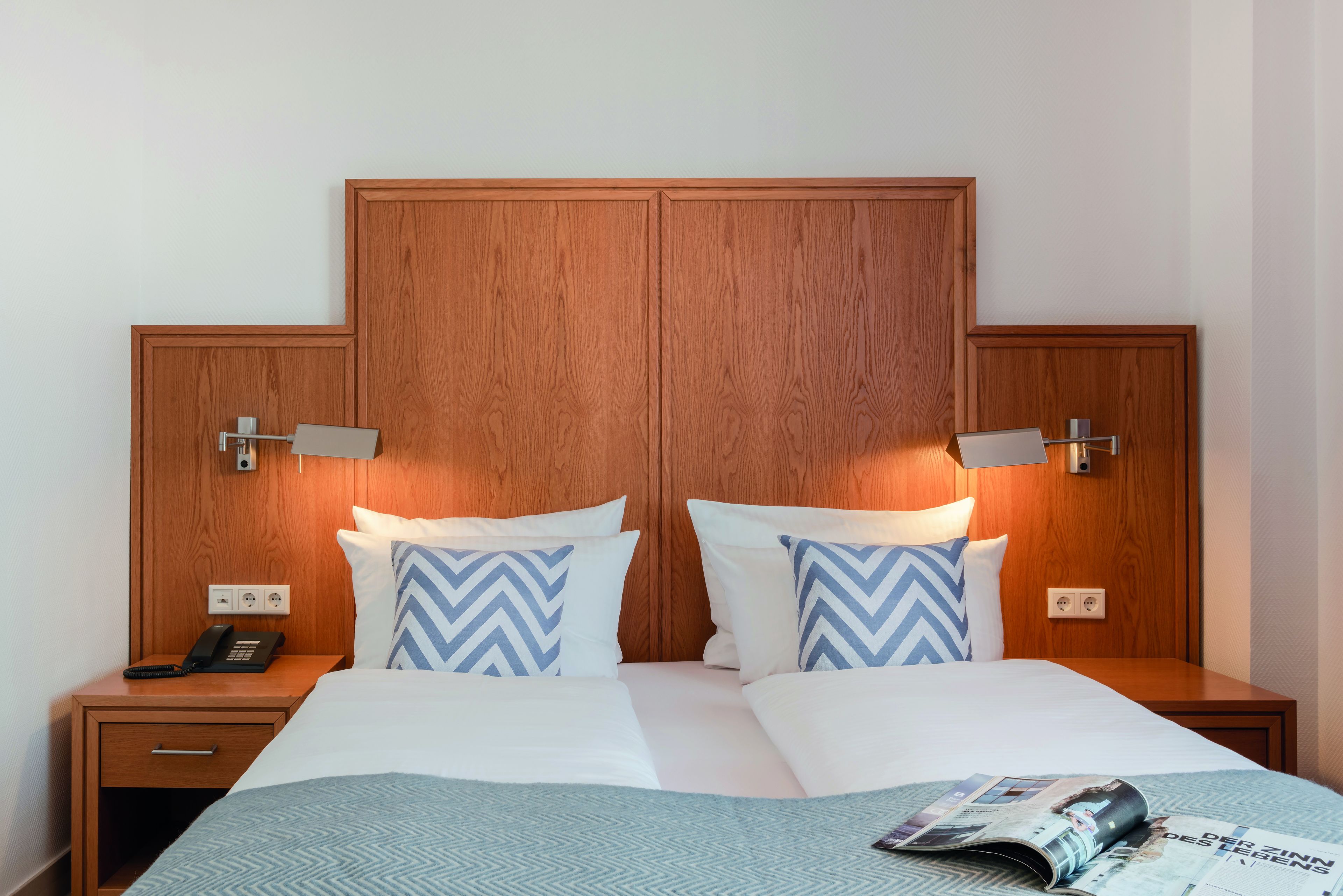 A hotel room with a wooden headboard, two bedside tables with lamps, a telephone, and a magazine on the bed.