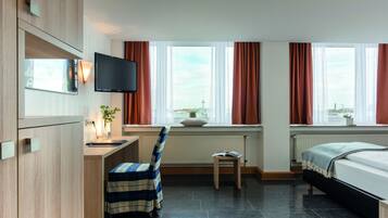 Habitación de hotel con cama, escritorio, silla, televisor y ventanas amplias con cortinas.