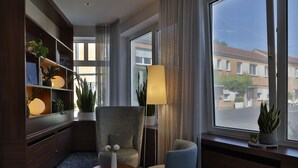 Une chambre d’hôtel moderne avec une grande fenêtre, un canapé, une petite table et une bibliothèque agrémentée de plantes.