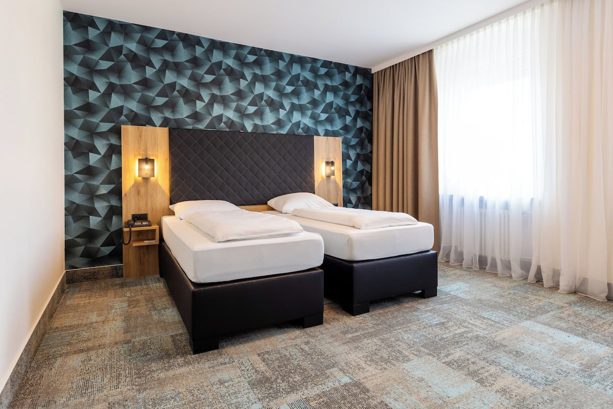 Et hotelværelse med moderne design, med væg i geometrisk mønster, en stor seng med træsenggavl og et stort vindue med lette gardiner.