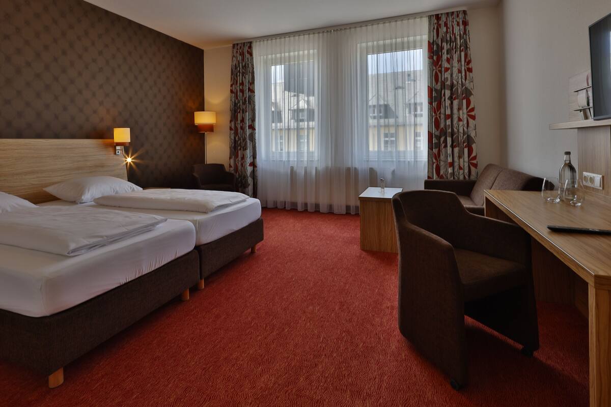 Et hotelværelse med to senge, et skrivebord, en stol, en lampe og et fjernsyn.