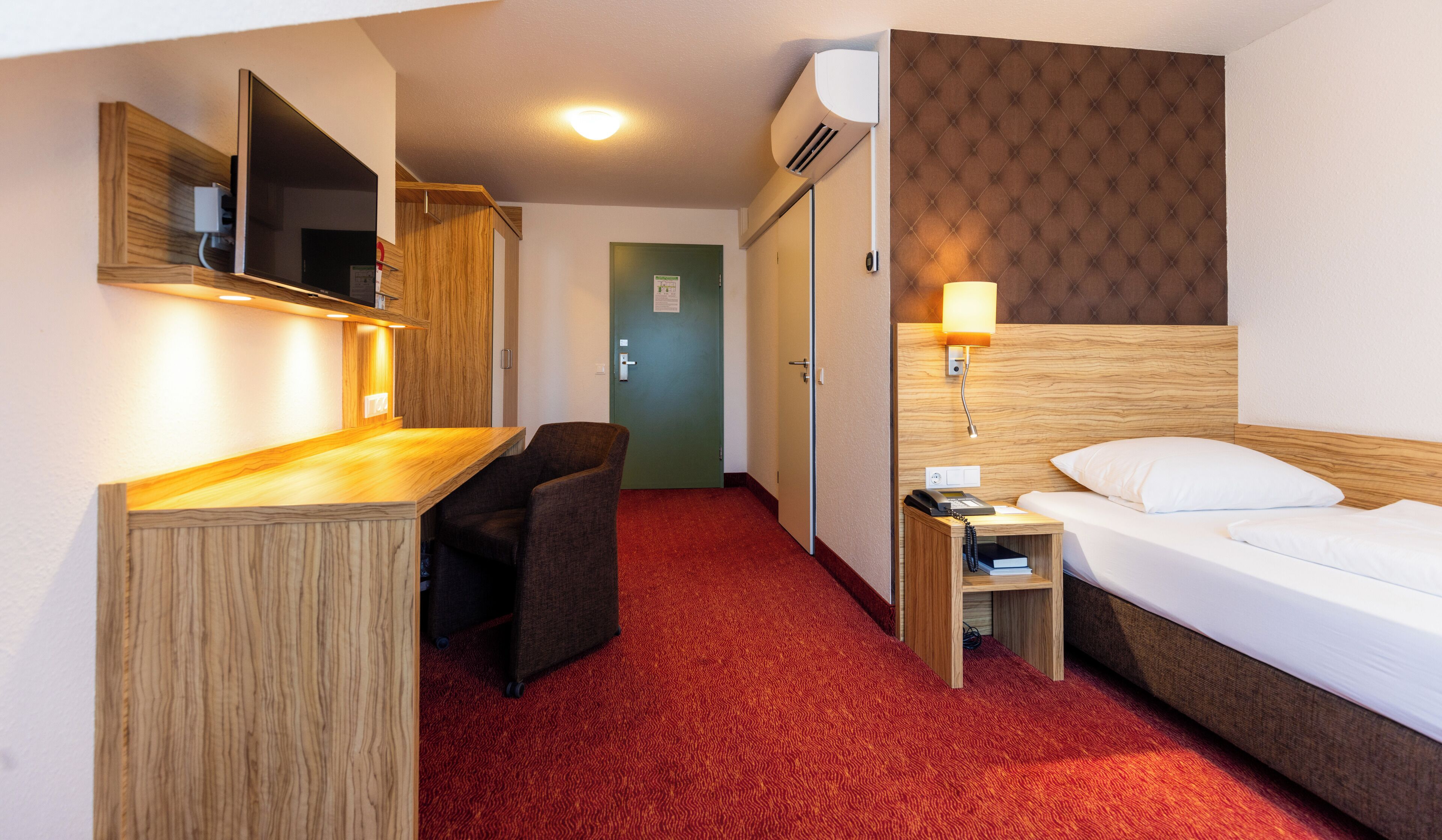 Ein Hotelzimmer mit einem Bett, einem Schreibtisch, einem Stuhl, einem Nachttisch, einer Lampe und einer Klimaanlage.