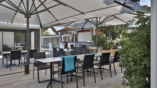 Un espace extérieur aménagé en terrasse, avec des tables et des chaises placées sous de grands parasols, le tout entouré de verdure.