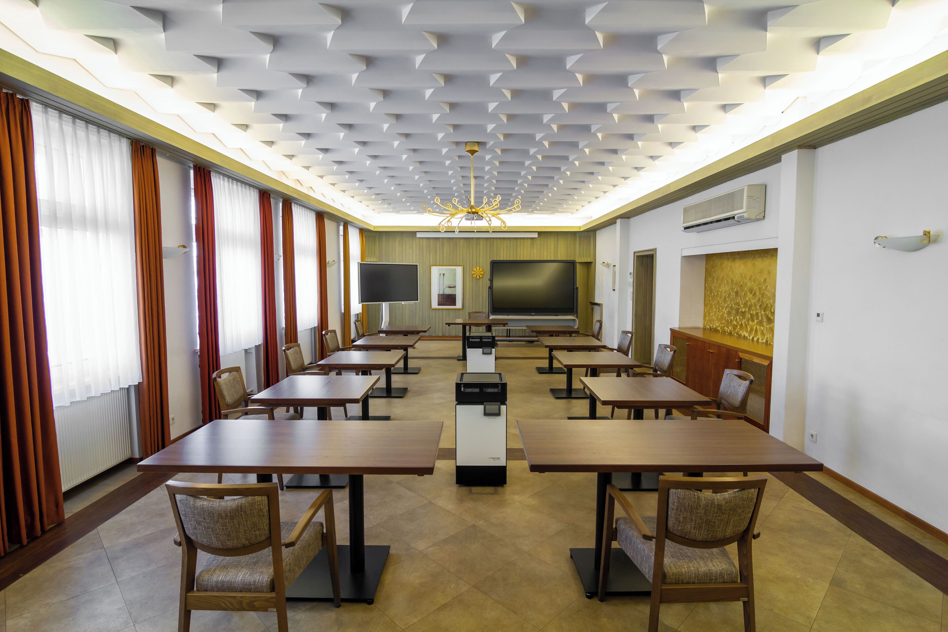 Un salón de conferencias con varias mesas y sillas, una pantalla grande y un techo con diseños geométricos.