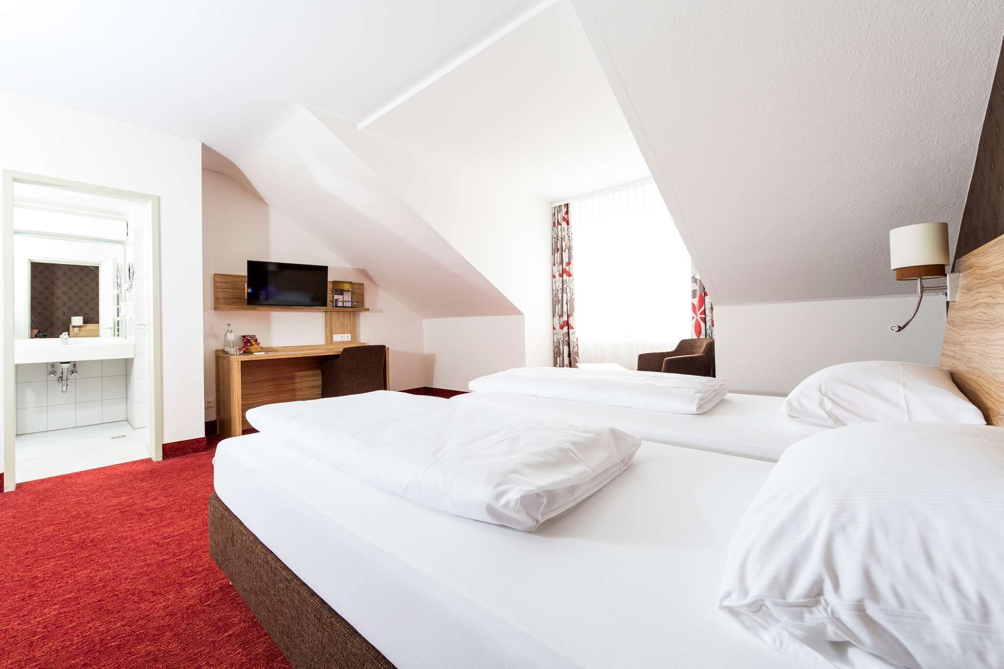 A hotel room with two beds, a desk with a TV, a bathroom, and a red carpet.