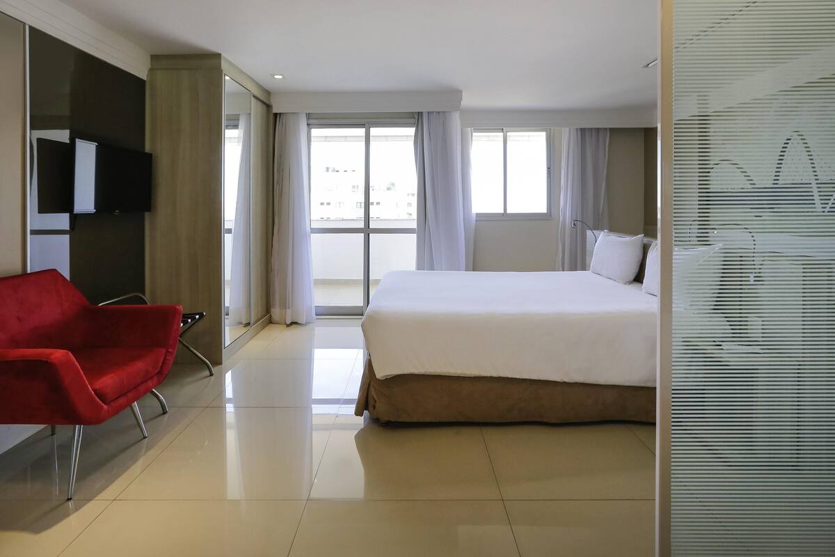 A hotel room with a large bed, a red chair, a flat-screen TV mounted on the wall, and a large window with curtains.
