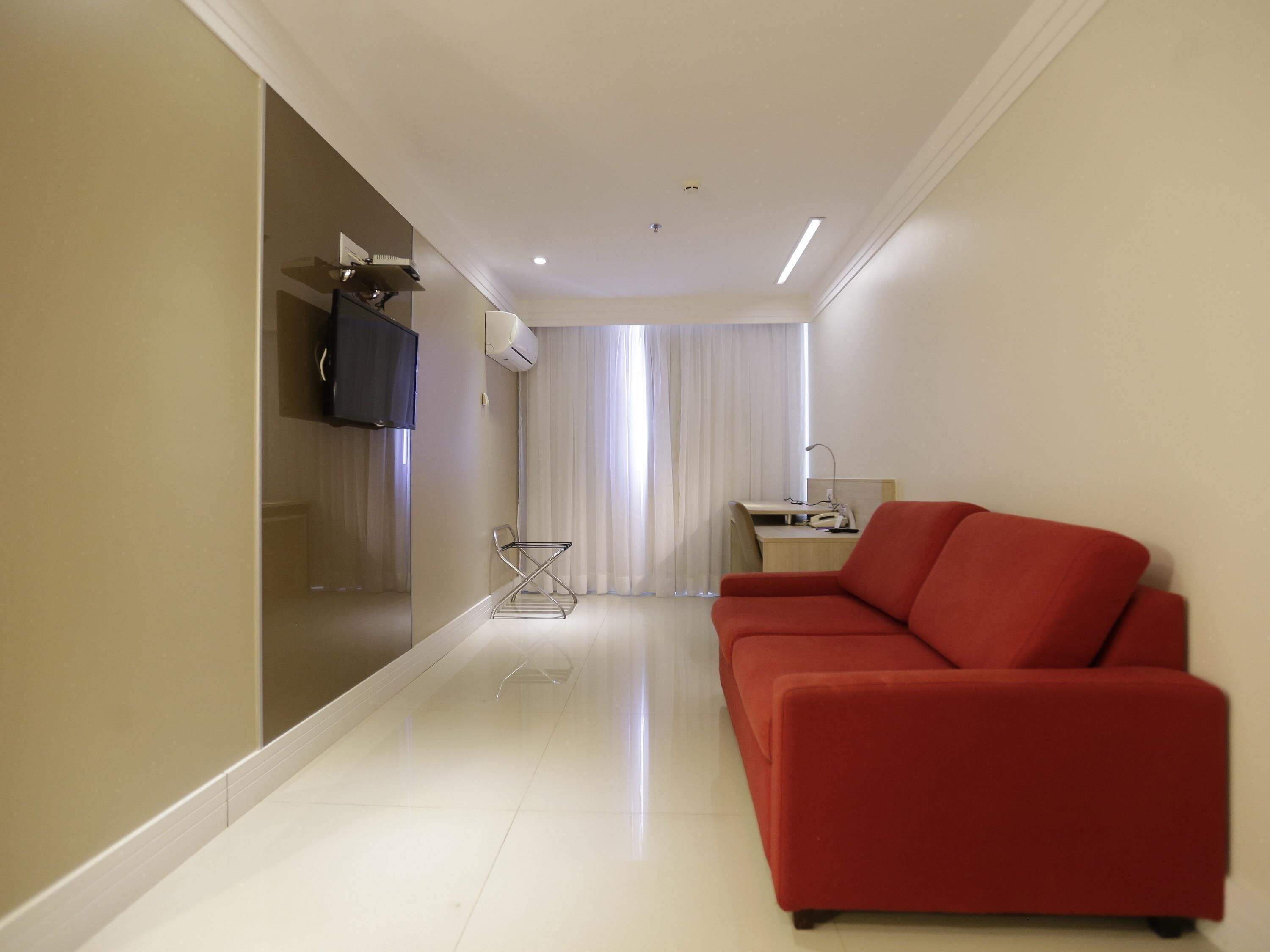A modern hotel room with a red sofa, a TV mounted on the wall, a desk with a chair, and a large window with curtains.
