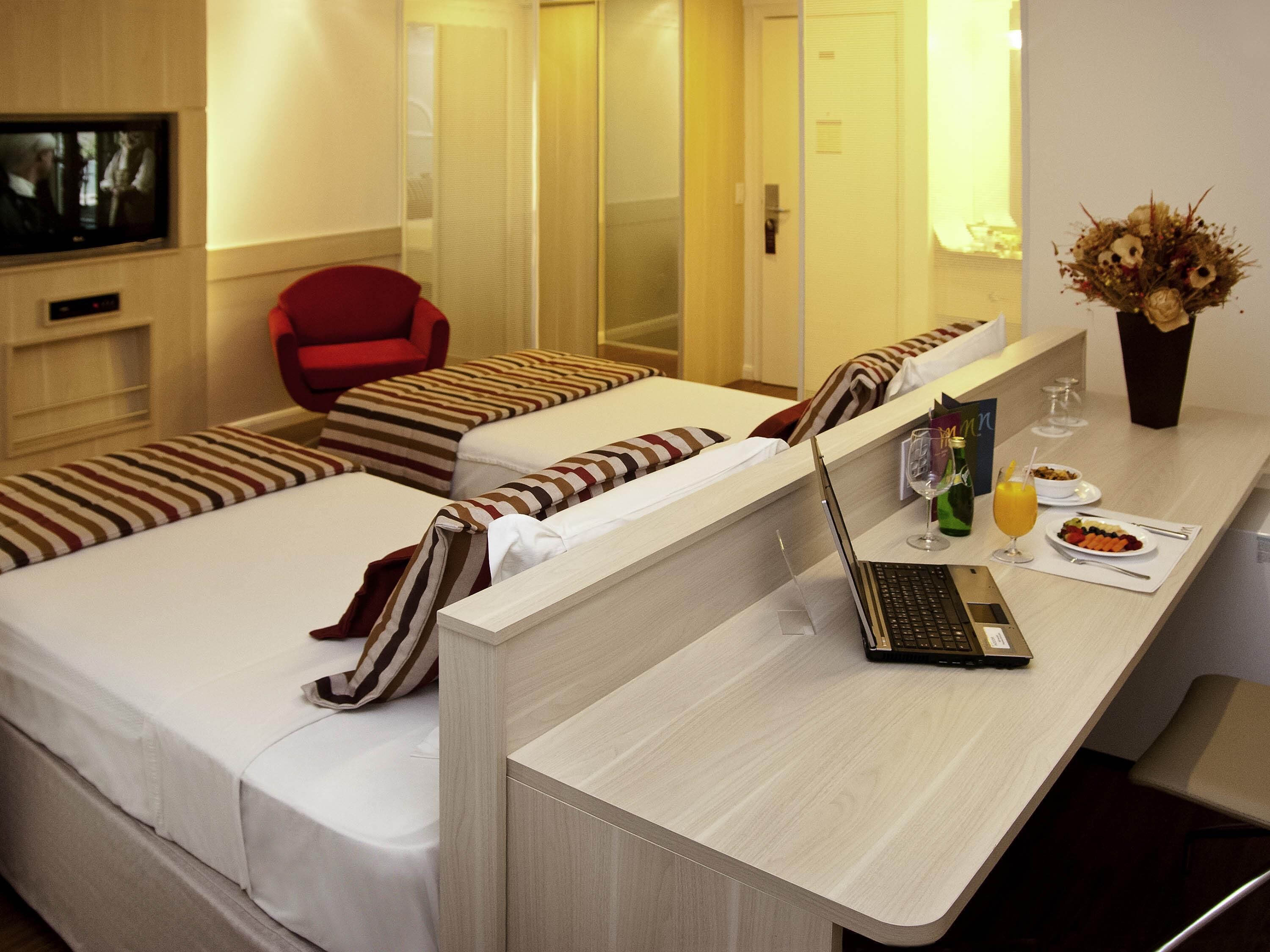 A hotel room with two single beds, a desk with a laptop, a red chair, a TV, and a vase with flowers.