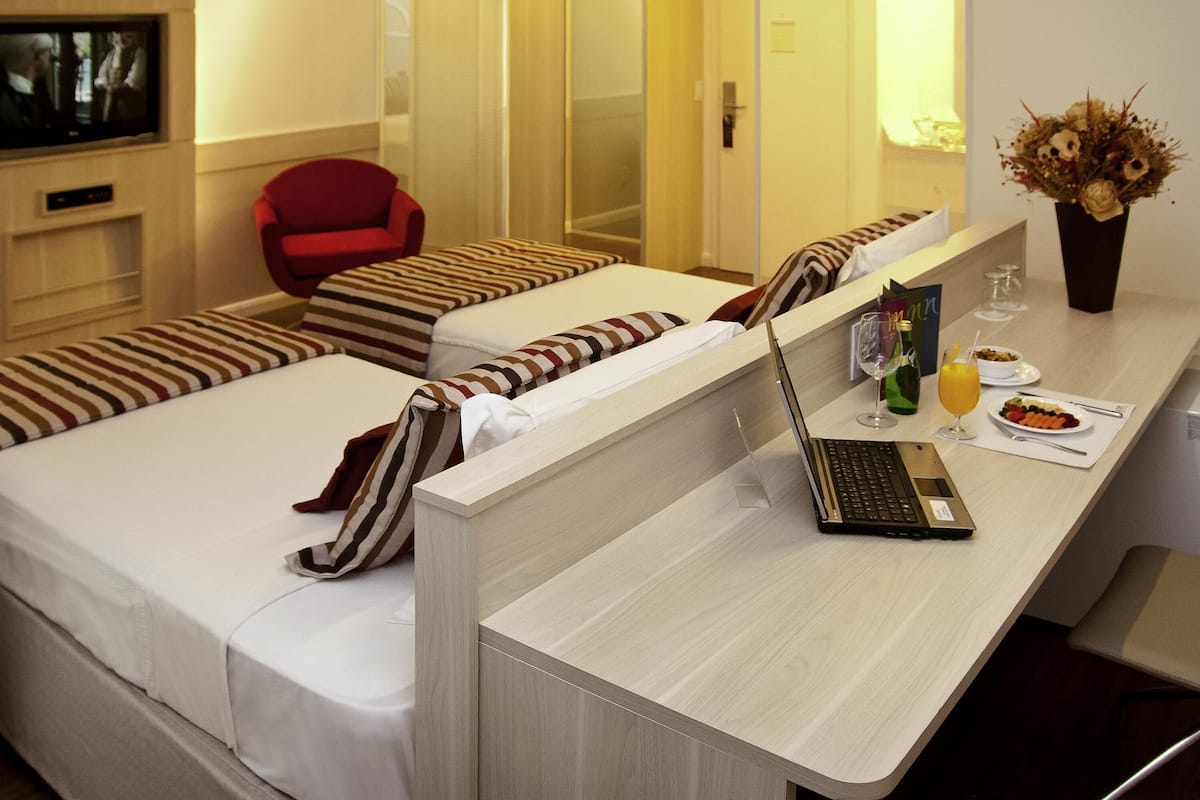Habitació d'hotel amb dos llits individuals, un escriptori amb un ordinador portàtil, una cadira vermella, una televisió i un ram de flors.