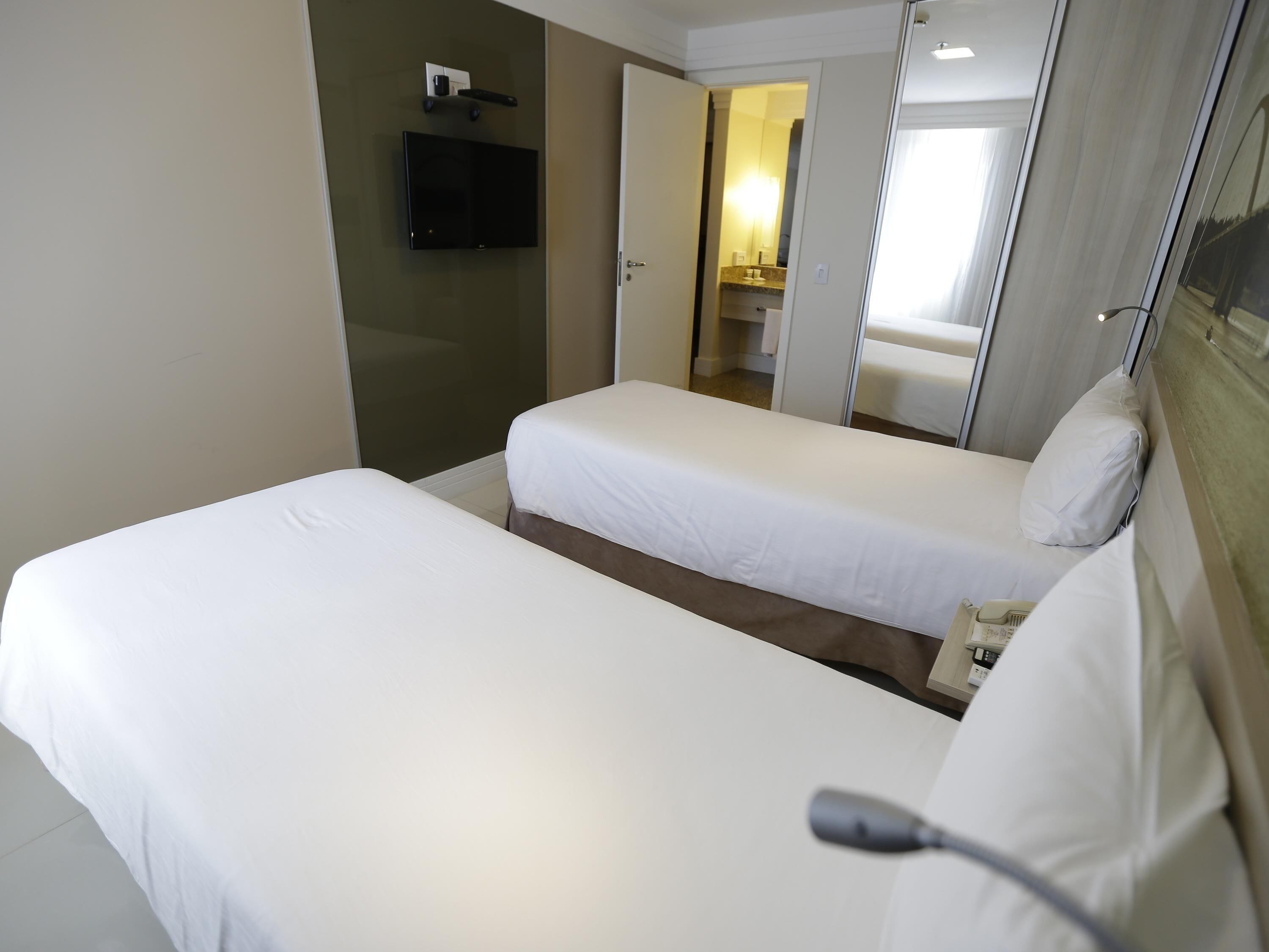 A hotel room with two single beds, a flat-screen TV mounted on the wall, and a bathroom visible through an open door.