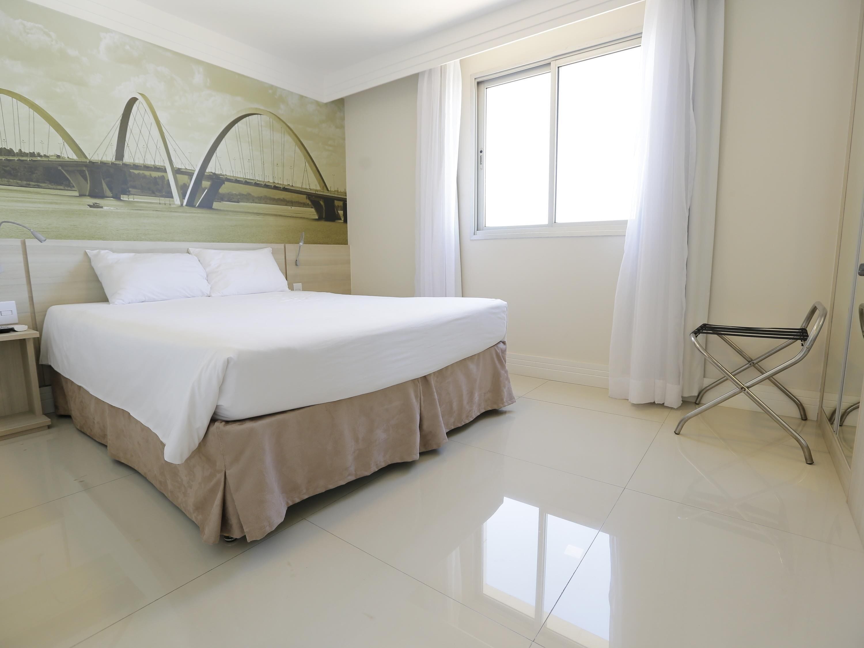 A hotel room with a large bed, a nightstand, a window with curtains, and a wall-mounted radiator.