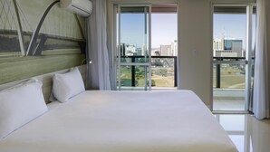Een groot bed met witte lakens in een hotelkamer met uitzicht op de stad door grote ramen.