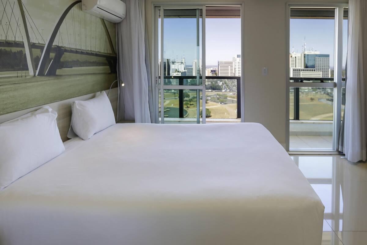 Una cama grande con ropa blanca en una habitación de hotel con vistas a la ciudad a través de amplios ventanales.
