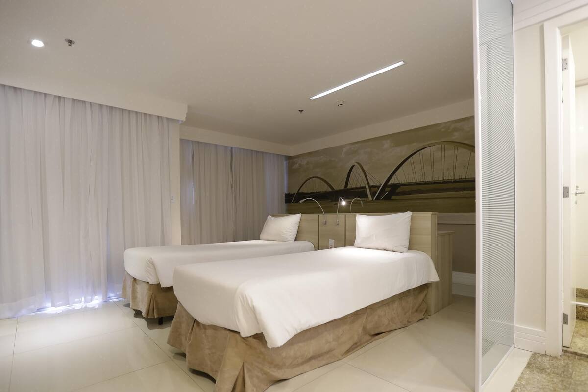 Habitación de hotel con dos camas, un ventanal grande con cortinas, una lámpara de techo moderna y una obra de arte colgada en la pared.